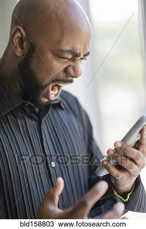 Stock Image - Frustrated black man using cell phone near window. Fotosearch