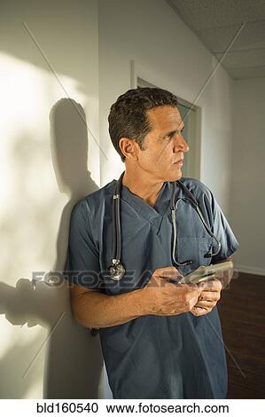 Stock Image - Hispanic doctor leaning on wall using cell phone. Fotosearch
