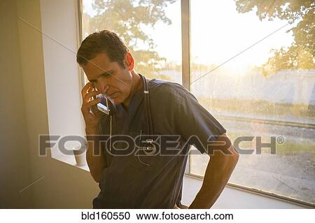 Stock Image - Hispanic doctor talking on cell phone at window. Fotosearch