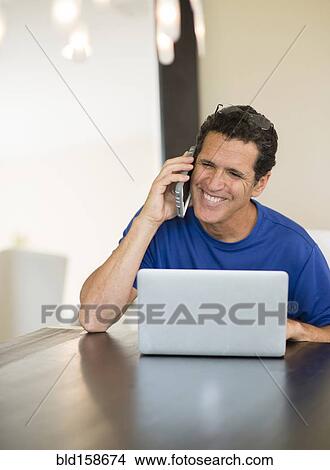 Picture - Hispanic man using laptop and cell phone at table. Fotosearch