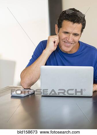 Stock Image - Hispanic man using laptop at table. Fotosearch