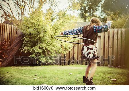 Stock Image - Mixed race girl in cowboy costume spinning plastic hoop in backyard. Fotosearch