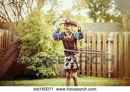 Stock Image - Mixed race girl in cowboy costume spinning plastic hoop in backyard. Fotosearch