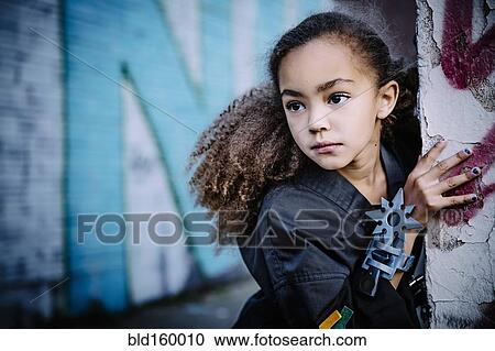 Stock Image - Mixed race girl in martial arts uniform with throwing star peeking around corner. Fotosearch