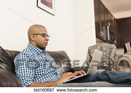 Stock Photo - Mixed race man using laptop on sofa in living room. Fotosearch