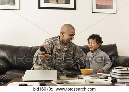 Stock Photography - Mixed race soldier father and son watching television in living room. Fotosearch