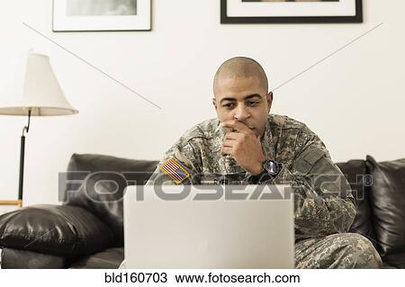 Stock Image - Mixed race soldier using laptop on living room sofa. Fotosearch