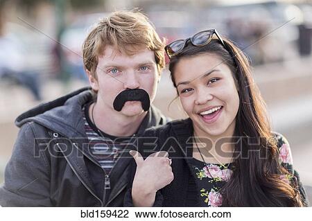 Stock Image - Teenage couple playing with fake mustache. Fotosearch