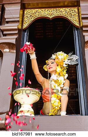 Stock Image - Asian woman scattering flowers from ornate temple window, Chiang Mai, Chiang Mai, Thailand. Fotosearch