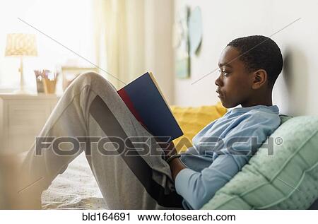 Stock Image - Black boy reading book on bed. Fotosearch