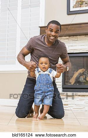 Picture - Black father helping baby son walk on living room floor. Fotosearch