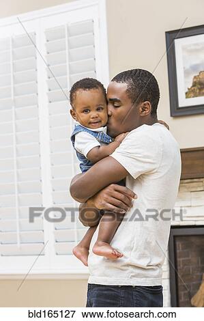 Stock Photo - Black father kissing baby son in living room. Fotosearch