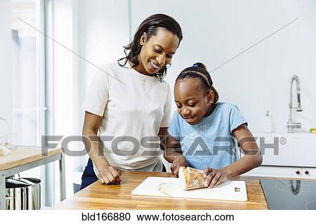 Stock Image - Black mother and daughter slicing bread in kitchen. Fotosearch