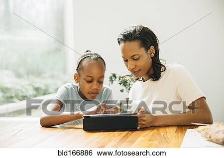 Stock Photograph - Black mother and daughter using digital tablet at table. Fotosearch