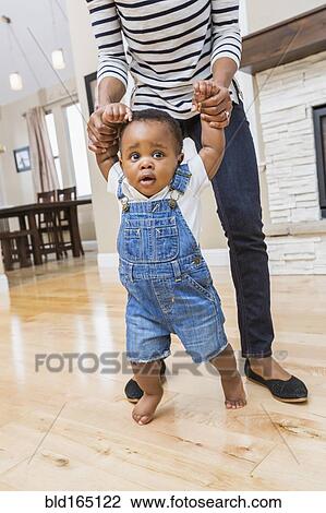 Stock Image - Black mother helping baby son walk on living room floor. Fotosearch