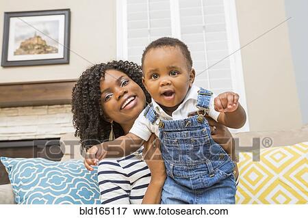 Stock Photo - Black mother holding baby son on sofa. Fotosearch
