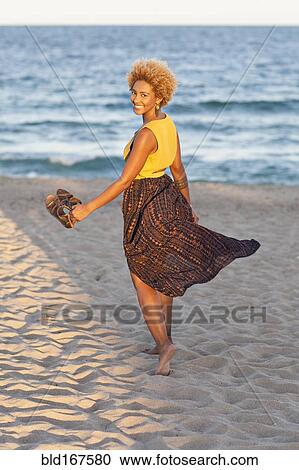 Stock Image - Black woman carrying sandals on beach. Fotosearch