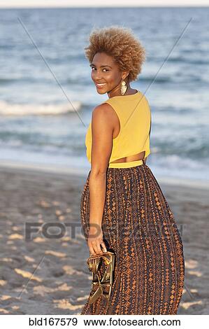 Stock Photo - Black woman carrying sandals on beach. Fotosearch
