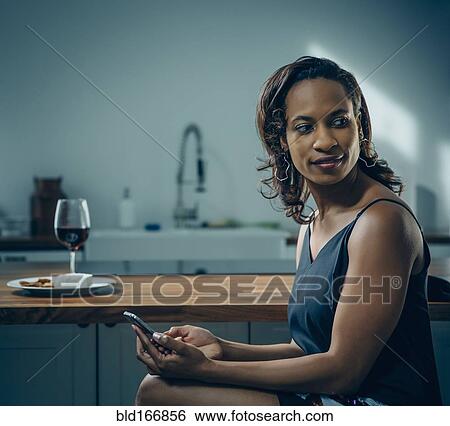 Stock Photograph - Black woman using cell phone at kitchen counter. Fotosearch