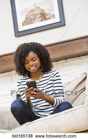 Stock Photo - Black woman using cell phone on sofa. Fotosearch