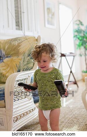 Stock Image - Caucasian baby boy carrying remote controls in living room. Fotosearch