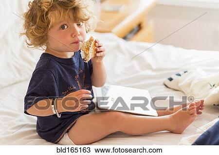 Stock Image - Caucasian baby boy eating and using digital tablet on bed. Fotosearch