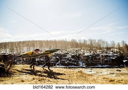 Stock Image - Caucasian couple carrying catamaran in snowy field. Fotosearch