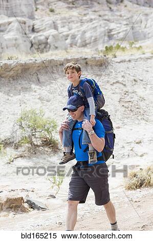Stock Photography - Caucasian father carrying son on shoulders on dirt path. Fotosearch