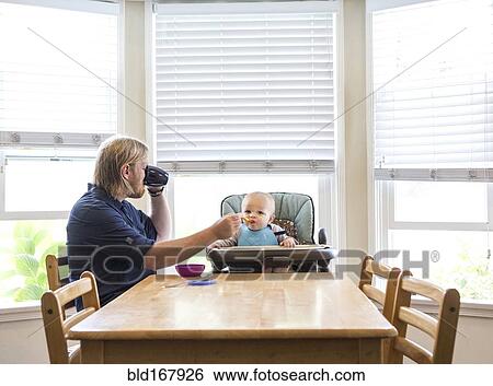Stock Photograph - Caucasian father feeding son in high chair. Fotosearch