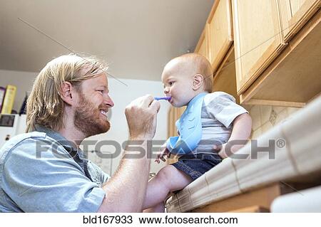 Stock Image - Caucasian father feeding son on kitchen counter. Fotosearch