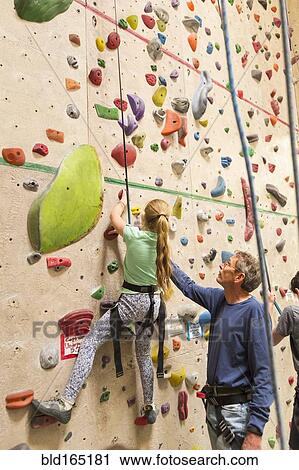Stock Image - Caucasian father helping daughter climb rock wall indoors. Fotosearch