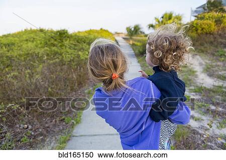 Stock Image - Caucasian girl carrying baby brother on wooden walkway. Fotosearch