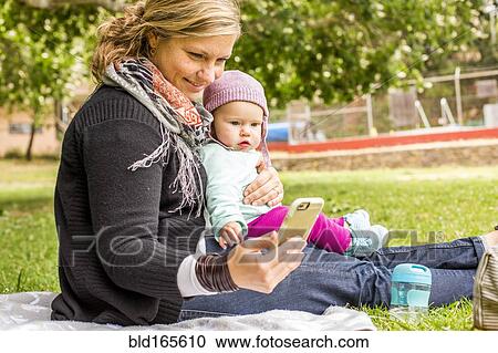 Stock Image - Caucasian mother and baby daughter using cell phone in park. Fotosearch