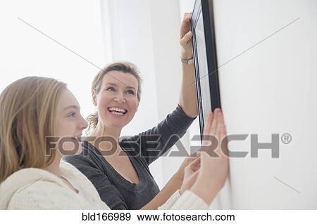Stock Photo - Caucasian mother and daughter hanging picture on wall. Fotosearch