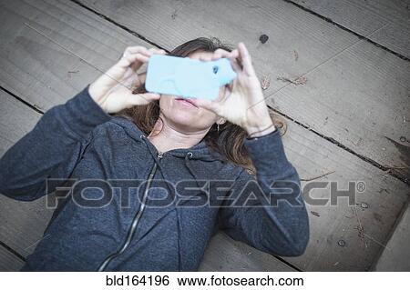 Stock Photograph - Caucasian woman laying on wooden deck taking cell phone selfie. Fotosearch