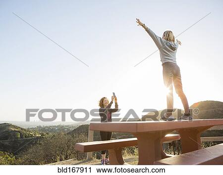 Stock Image - Caucasian woman photographing friend on hilltop. Fotosearch