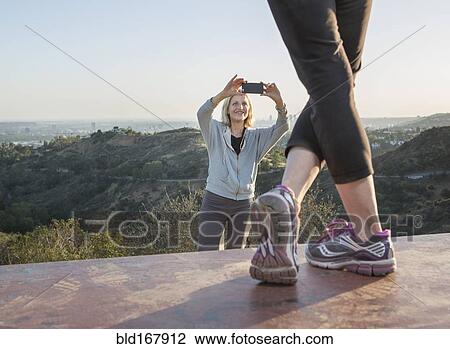 Stock Image - Caucasian woman photographing friend on hilltop. Fotosearch