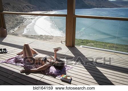 Stock Photo - Caucasian woman sunbathing and reading book on deck over beach. Fotosearch