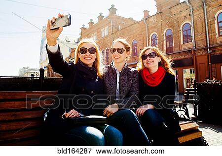 Stock Photo - Caucasian women taking cell phone photographs on city bench. Fotosearch