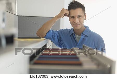 Stock Image - Chinese businessman smiling near filing cabinet. Fotosearch