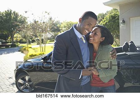 Picture - Couple hugging in driveway at convertible. Fotosearch