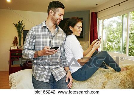 Couple using cell phones on sofa in living room View Large Photo Image Stock Image - Couple using cell phones on sofa in living room. Fotosearch