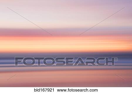 Stock Image - Defocused view of ocean waves on beach under sunset sky. Fotosearch