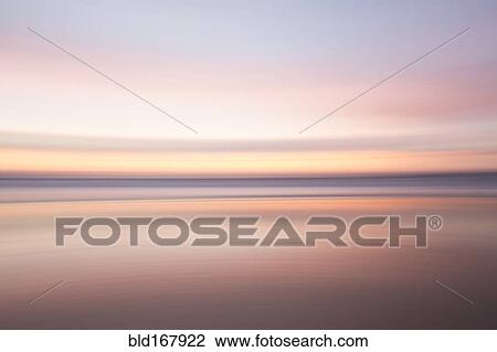 Stock Image - Defocused view of ocean waves on beach under sunset sky. Fotosearch