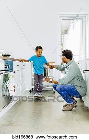 Stock Photo - Father helping son balance on skateboard in kitchen. Fotosearch