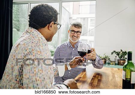 Stock Image - Gay couple toasting with wine at counter. Fotosearch