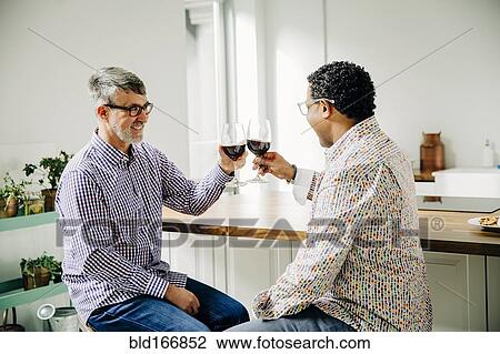 Stock Image - Gay couple toasting with wine at counter. Fotosearch