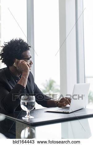 Stock Image - Hispanic businessman using cell phone and laptop in office. Fotosearch