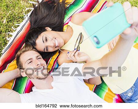 Stock Image - Hispanic couple taking selfie on blanket in park. Fotosearch