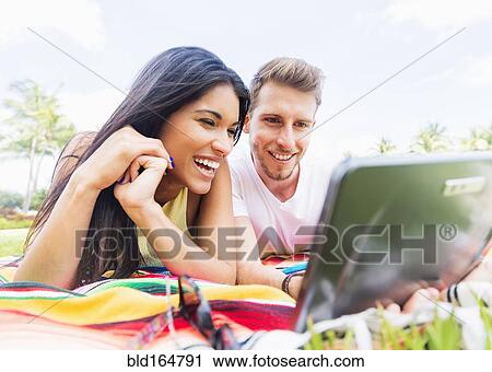 Stock Image - Hispanic couple using digital tablet on blanket in park. Fotosearch